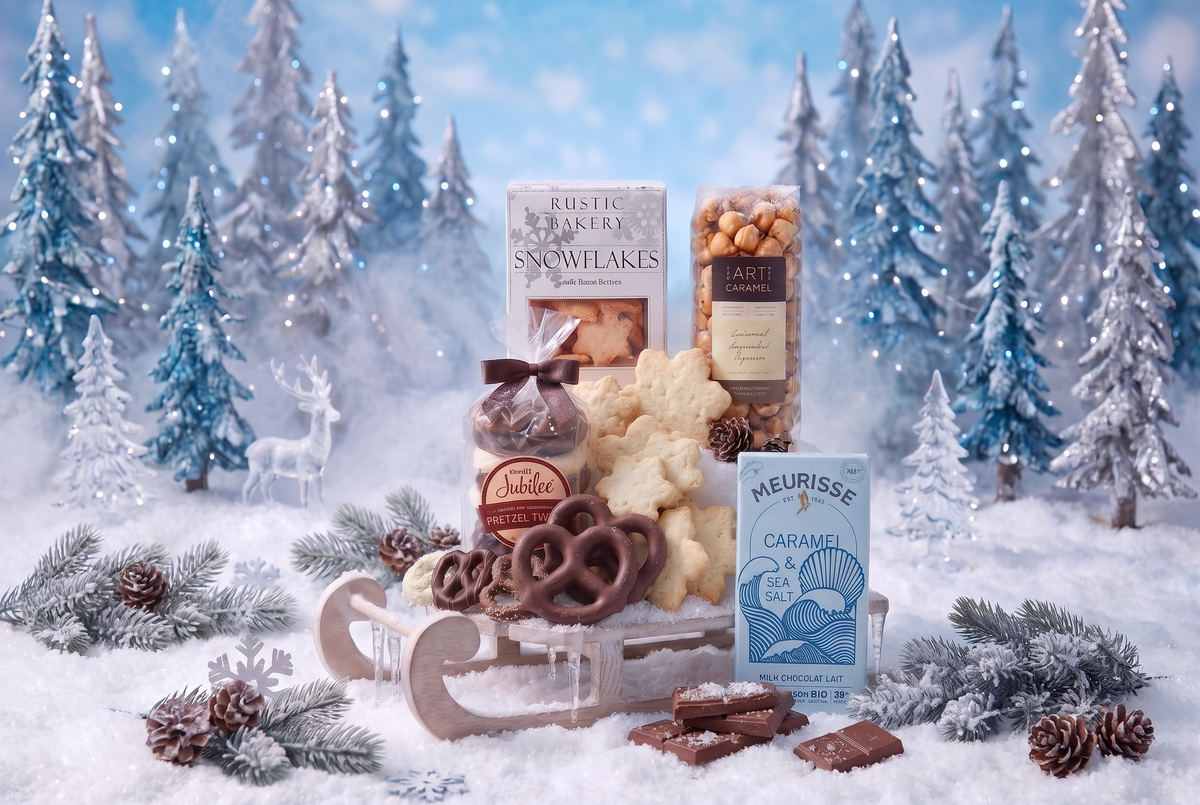 Holiday gourmet treats with snowflake cookies, chocolate-covered pretzels, caramel nuts and a chocolate bar arranged on a miniature sleigh among snowy trees.