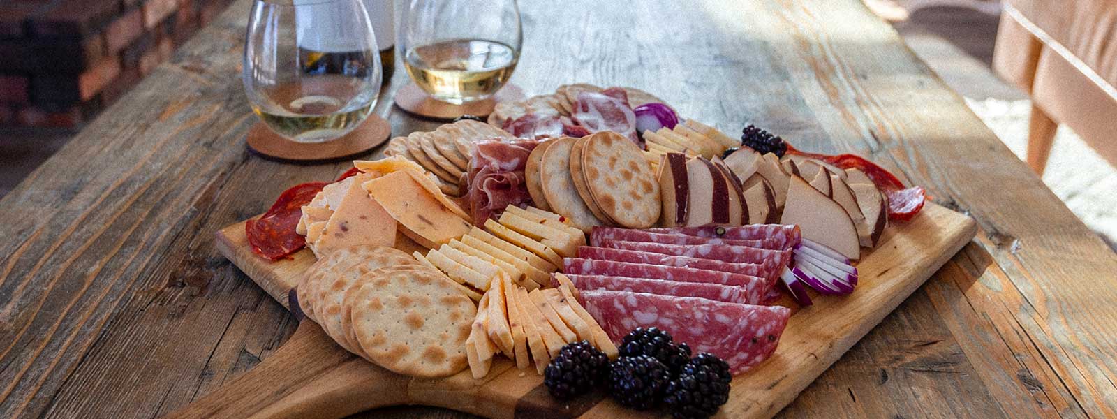 Housewarming gift display on a wooden table featuring a charcuterie platter and two beverages, ideal for welcoming someone to a new home.