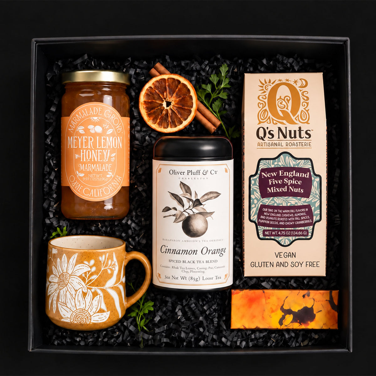 BLACK GIFT BOX WITH MEYER LEMON HONEY MARMALADE, CINNAMON ORANGE TEA, A PACKAGE OF GOURMET NUTS, A CHOCOLATE ORANGE CHOCOLATE BAR AND A CUP WITH SUNFLOWERS -