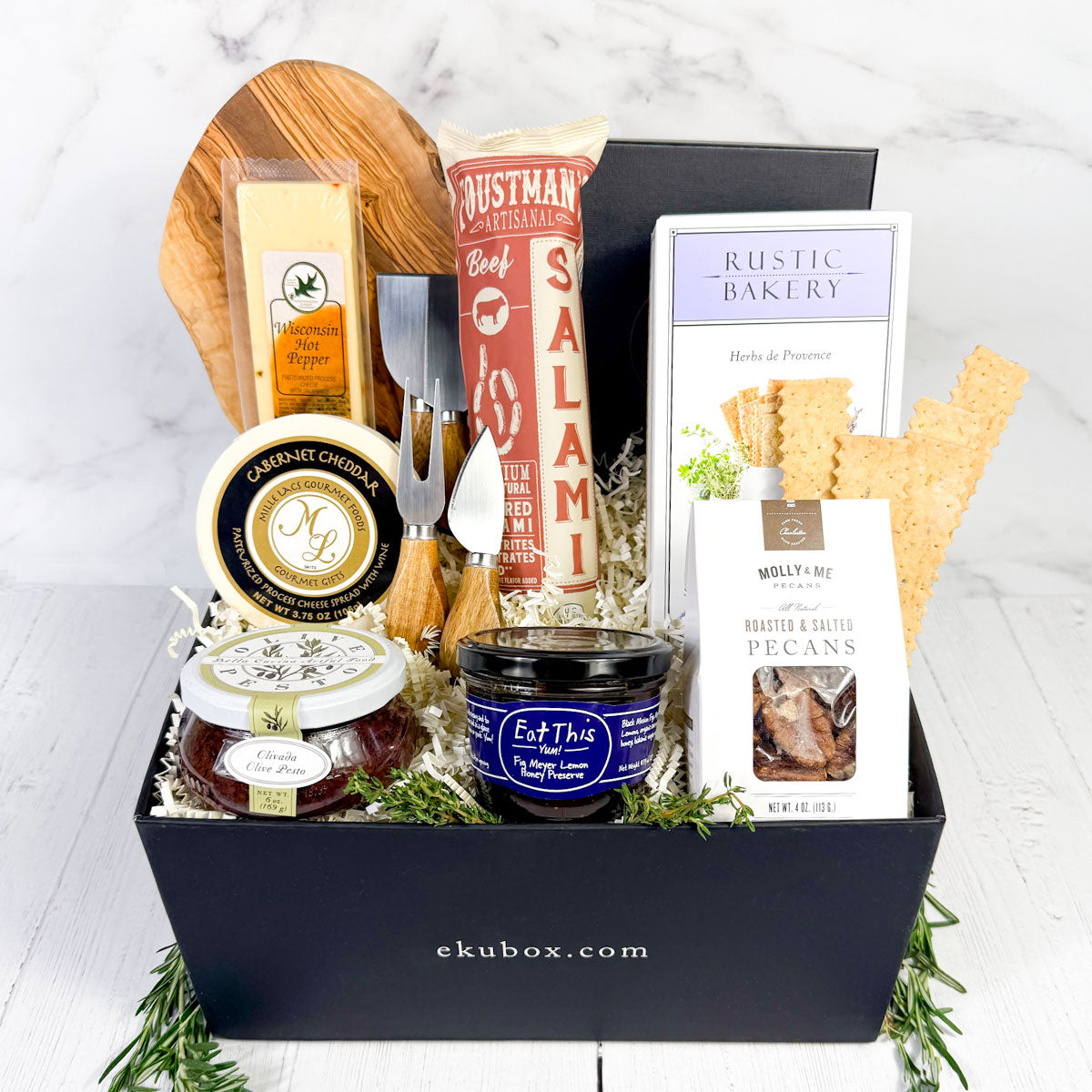 A charcuterie gift set displayed in a black box, including a wood cutting board, a 4-piece acacia wood handle utensil set, various gourmet cheeses, salami, pesto, honey, and crackers.
