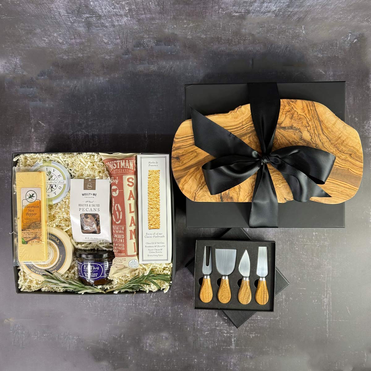 top view of A charcuterie gift set displayed in a black box, including a wood cutting board, a 4-piece acacia wood handle utensil set, various gourmet cheeses, salami, pesto, honey, and crackers.
