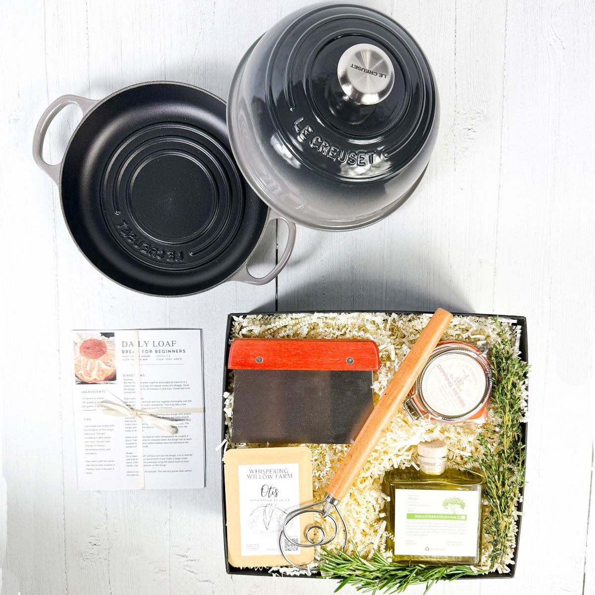 A Le Creuset bread baking kit with a grey bread oven, dutch bread whisk, sourdough starter, bench scraper, and olive oil with herbs, on a white marble surface.