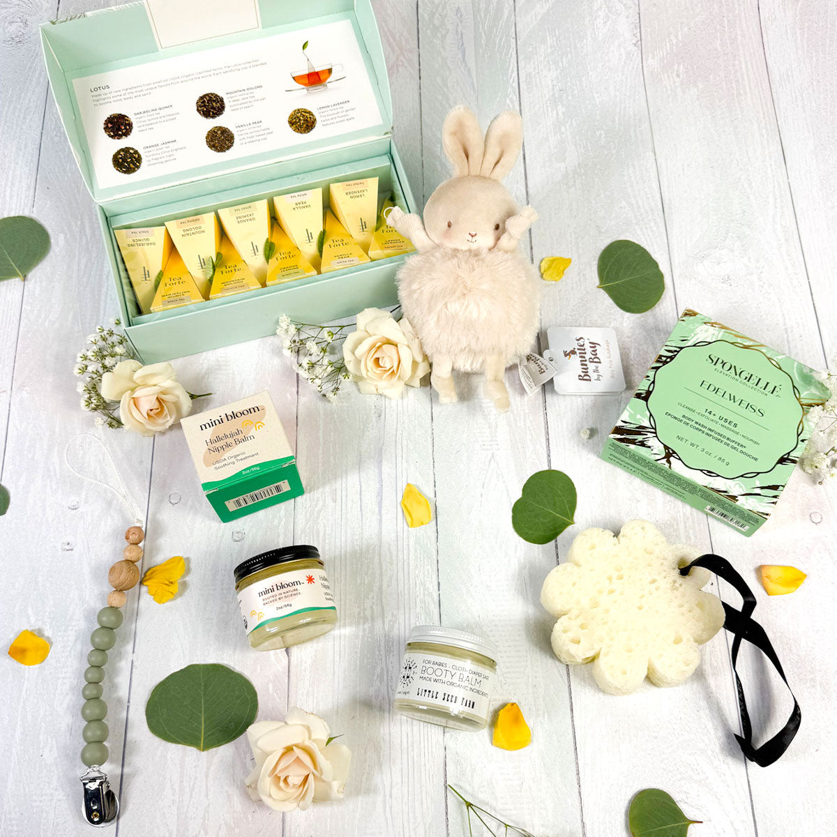 Mom and Baby gift box with nipple balm, Tea Forté, Spongellé body buffer, plush baby toy, goat milk baby cream and a silicone bead and wood pacifier clip on a white wood background