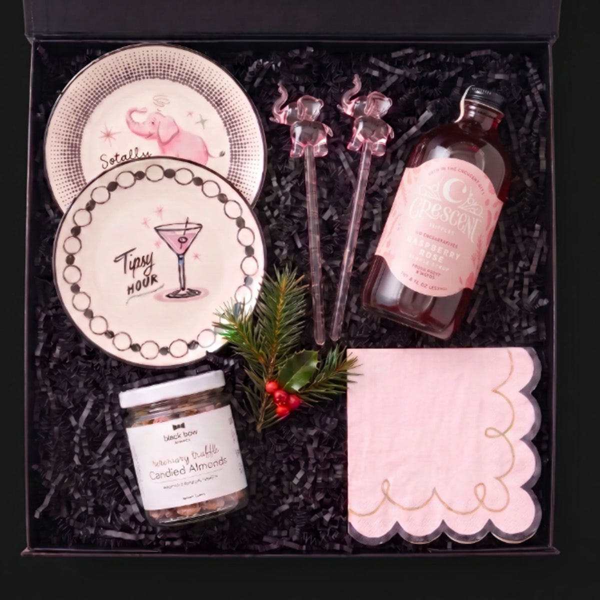 A curated gift box filled with black shredded paper, featuring a pink-and-white elephant-patterned plate, a martini-themed plate, a bottle of non-alcoholic mixer, two pink elephant-shaped stir sticks, a jar of candied almonds, and a pink napkin with gold script.
