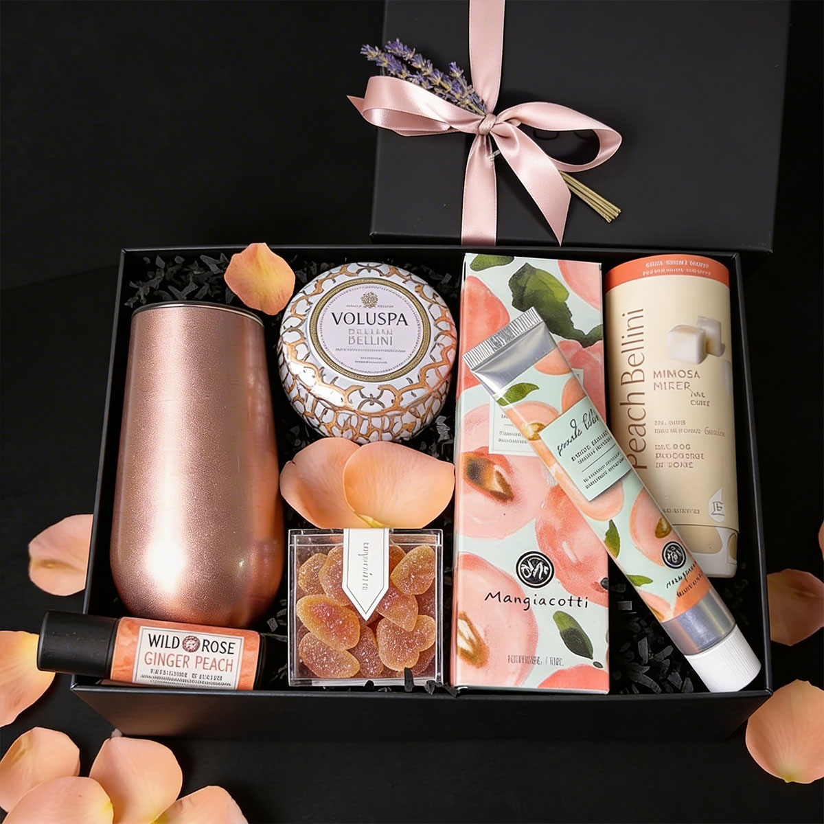 A luxury black ekubox gift set containing a rose gold thermal flute, a Voluspa candle, Sugarfina gummies, peach hand cream, Teaspressa bellini sugar cubes, and lip balm, styled with peach rose petals.