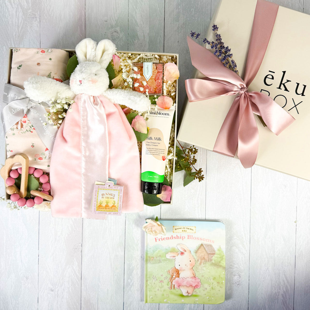 Baby girl gift box with a pink bunny lovey, floral swaddle, baby book, teether, and body cream with box top on white background.