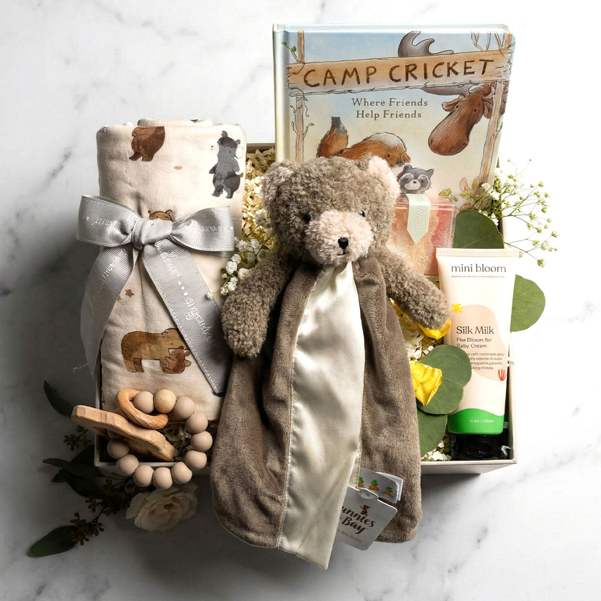 A gender-neutral baby gift box featuring a plush bear lovey, swaddle blanket, teether, baby cream, and Sugarfina gummies. on white marble background
