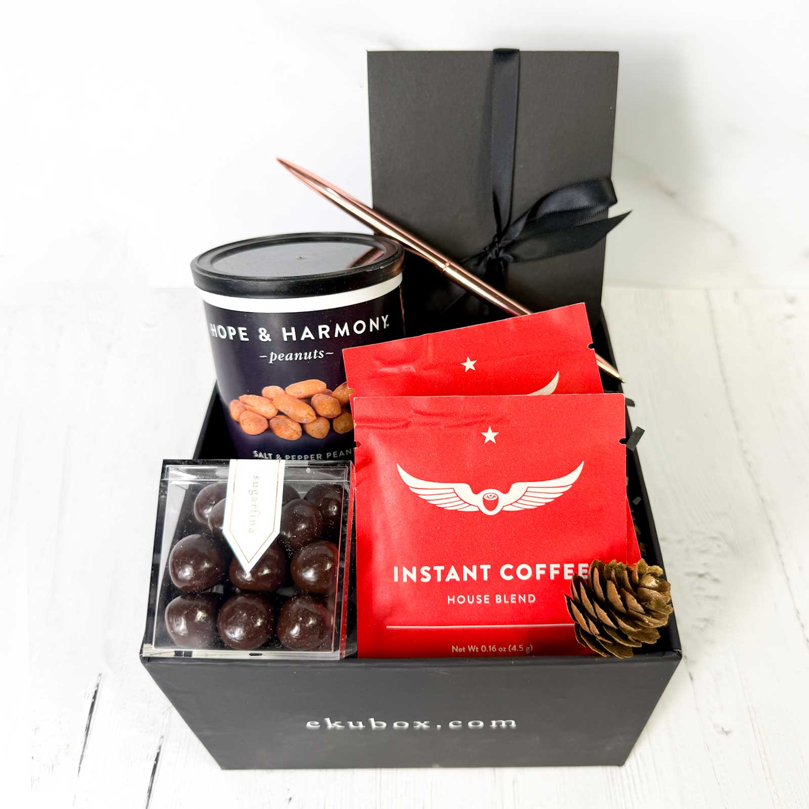 Gift set in a black box with instant gourmet coffee, snacks, and a rose gold pen on a white surface