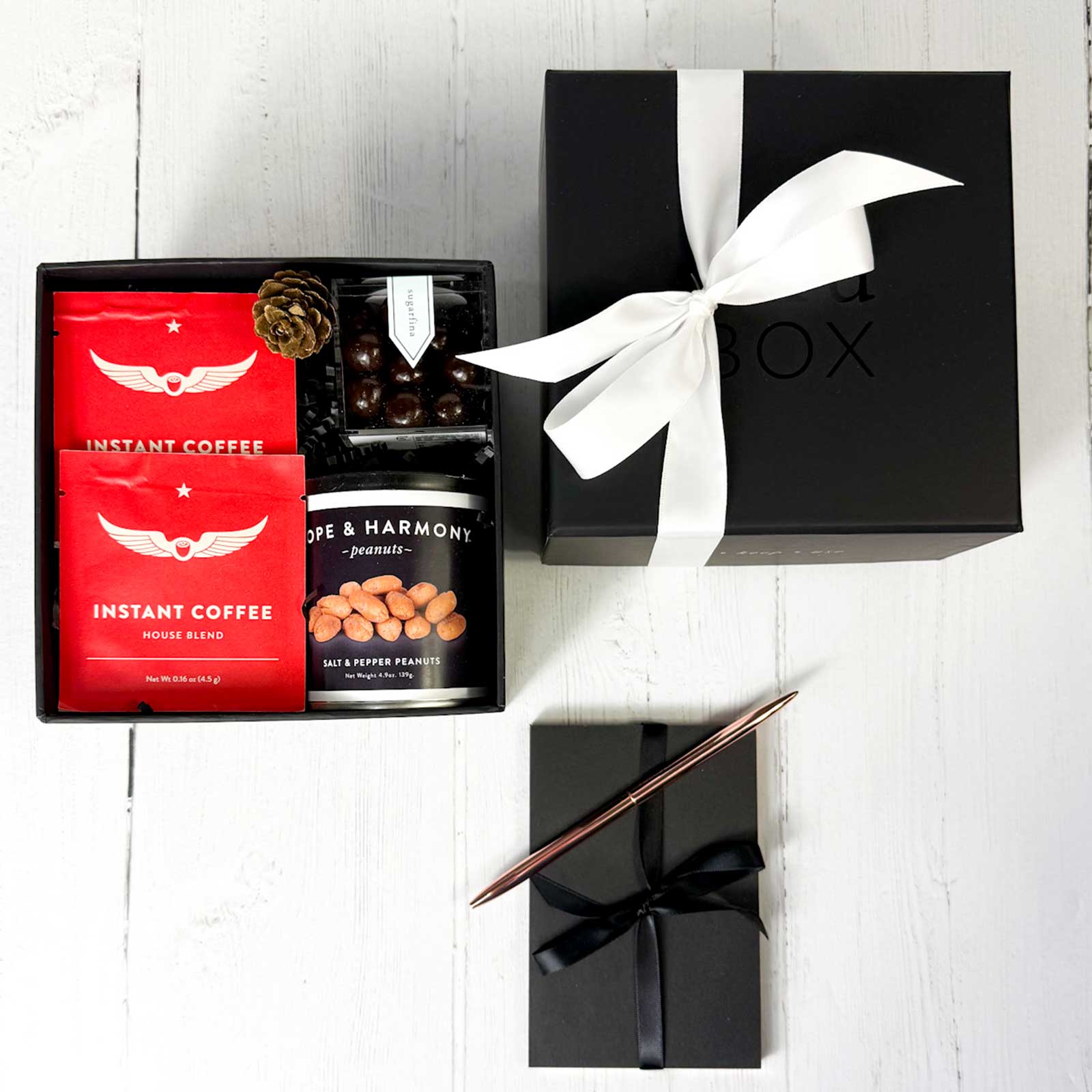 Gift set in a black box with instant gourmet coffee, snacks, and a rose gold pen on a white surface