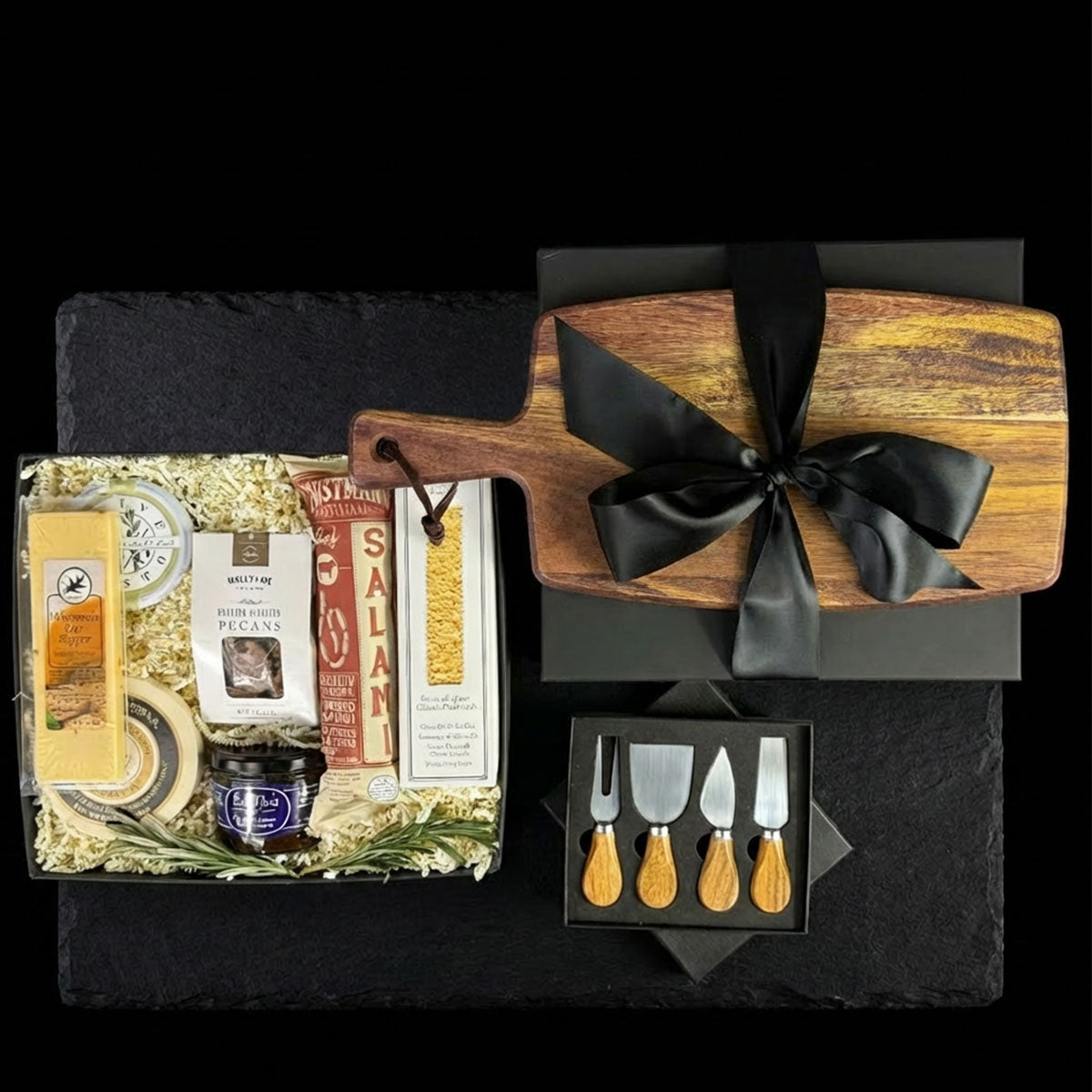 charcuterie gift set displayed in a black box, including a wood cutting board, a 4-piece acacia wood handle utensil set, various gourmet cheeses, salami, pesto, honey, and crackers.