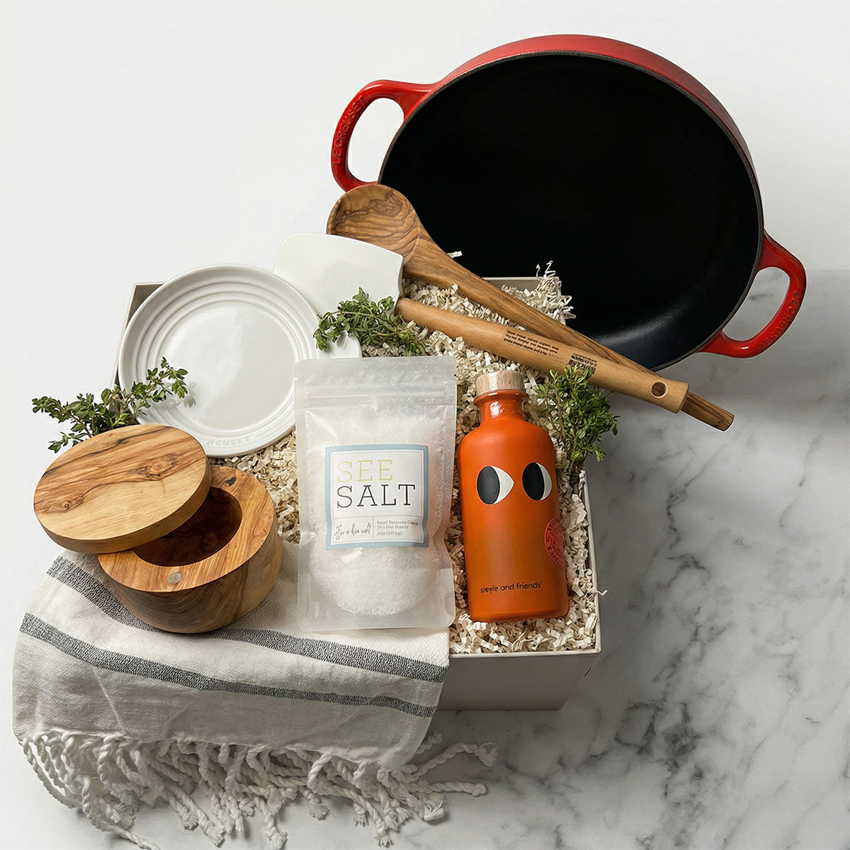 Le Creuset Signature Everyday Pan gift set on a marble surface with a white spoon rest, olive wood spoon and salt cellar, towel, and olive oil.