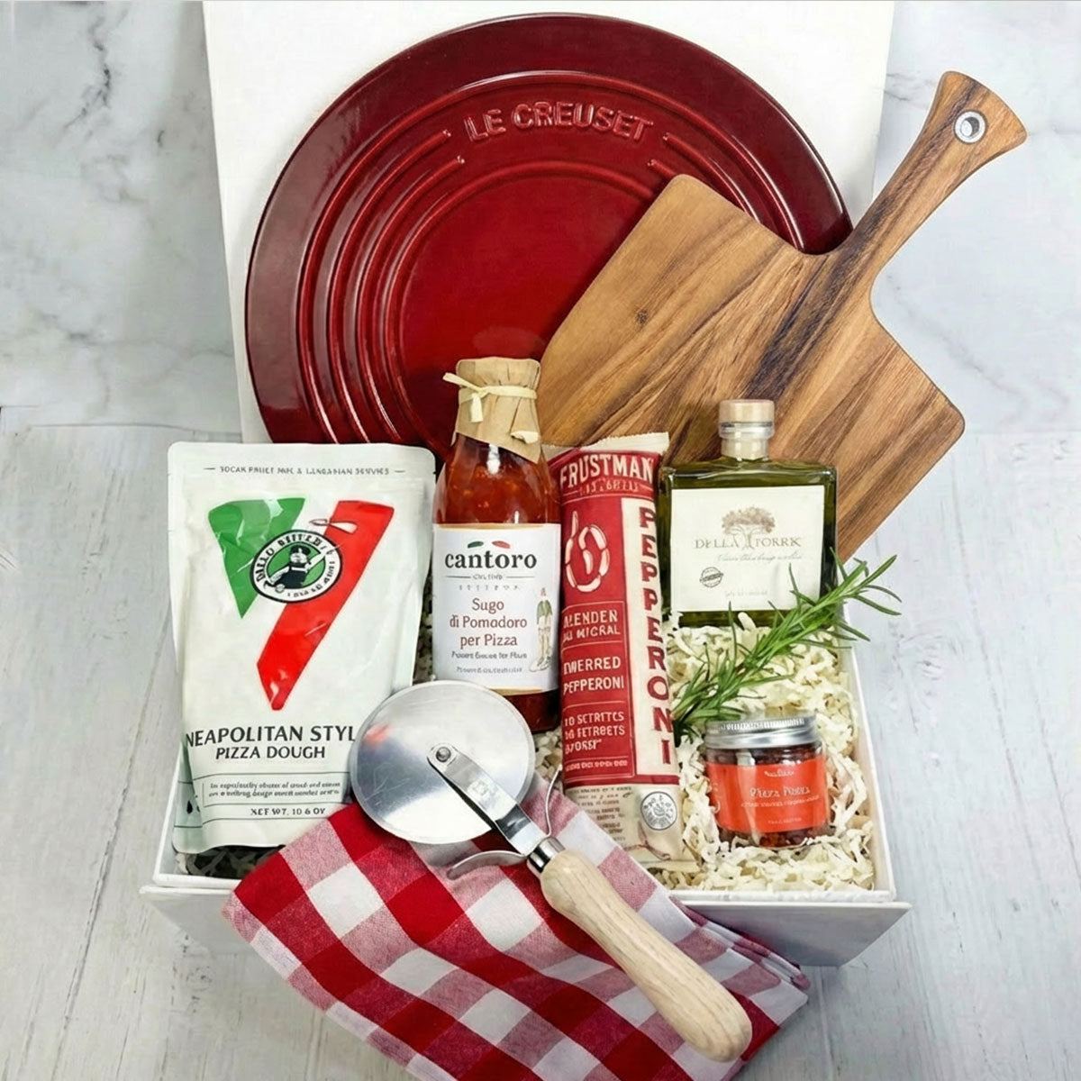 Gourmet Pizza Party box with pizza-making items including a red Le Creuset dish, pizza dough, and tools on a white surface.