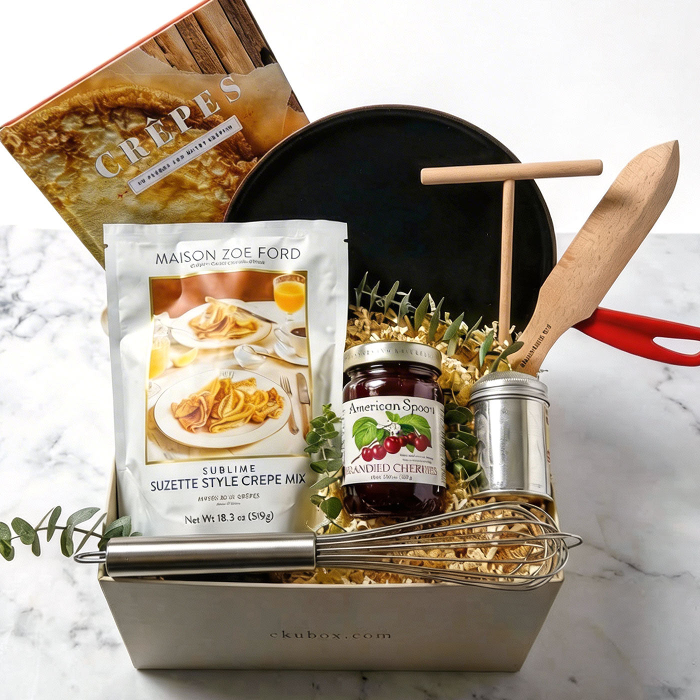 An open luxury gift box containing a black Le Creuset enameled cast iron crepe pan, a jar of American Spoon brandied cherries, Maison Zoe Ford crepe mix, a stainless steel whisk, a sugar sifter, and wooden crepe tools,