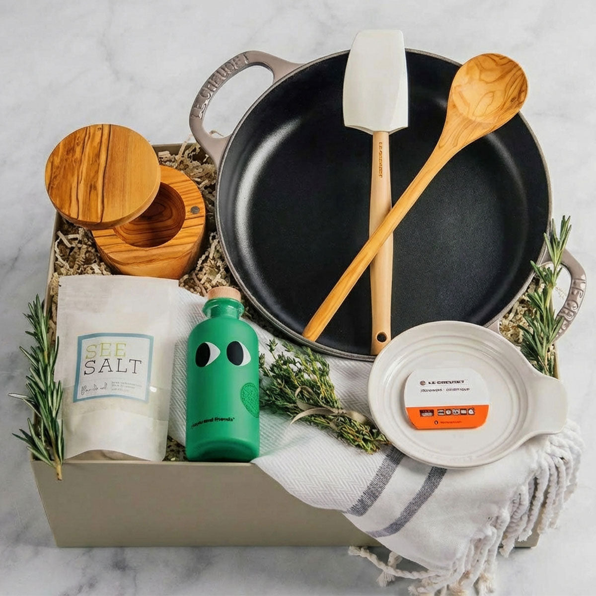 Le Creuset Signature Everyday Pan gift set on a marble surface with a white spoon rest, olive wood spoon and salt cellar, Turkish Fouta towel, Fleur de Sel, and Greek olive oil.