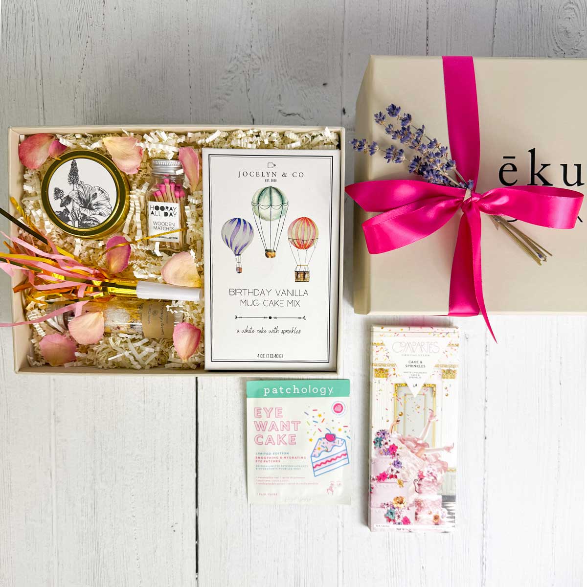 top view of Birthday gift box featuring cake-themed treats and self-care items, including a vanilla mug cake mix, sprinkle chocolate bar, candle, colorful matches, eye patches, floral bath salts, and a party blow horn arranged in a small gift box.