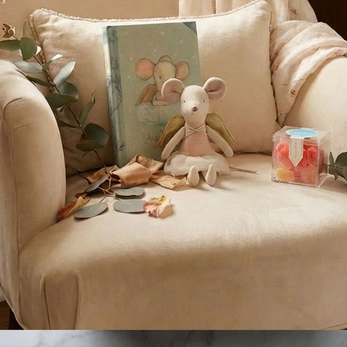 Stuffed maileg mouse toy on a beige chair with a children's book and cube of candy