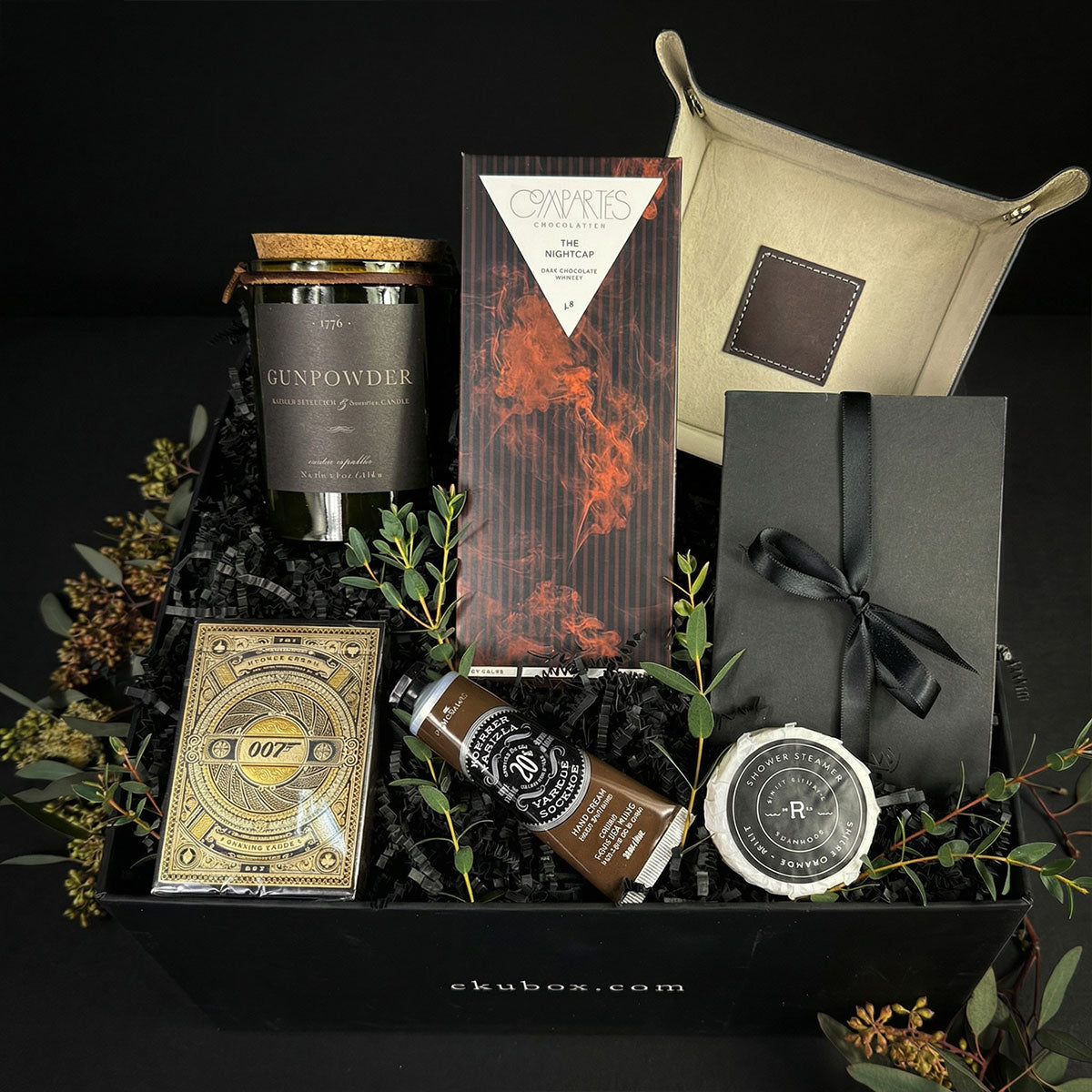 A men's luxury gift box containing a brown leather snap valet tray, Theory11 playing cards, a notebook, chocolate bar, scented candle, hand cream, and a shower steamer.