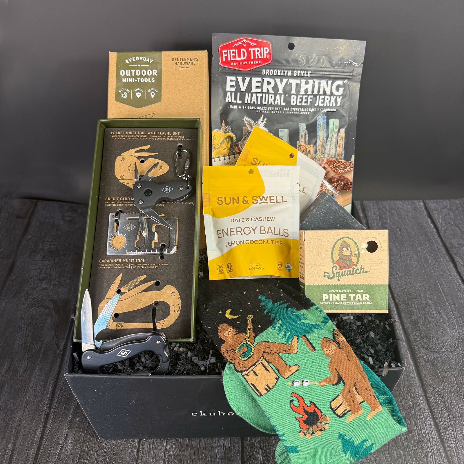 A collection of outdoor-themed items including a Sasquatch-themed sock, a pine tar bar of soap, a mini toolkit with a multi-tool with a flashlight, screwdriver, and other tools, a package of gourmet jerky, and a package of organic trail mix, all displayed in a black gift box.