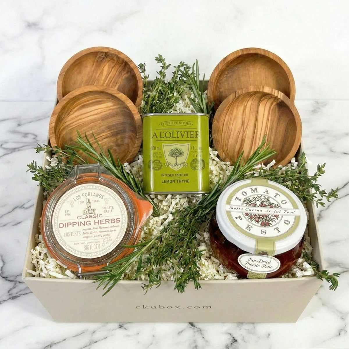 A curated gift box featuring a bottle of dipping herbs, a jar of tomato pesto, olive oil and olive wood dipping bowls arranged with rosemary sprigs on a white marble surface.