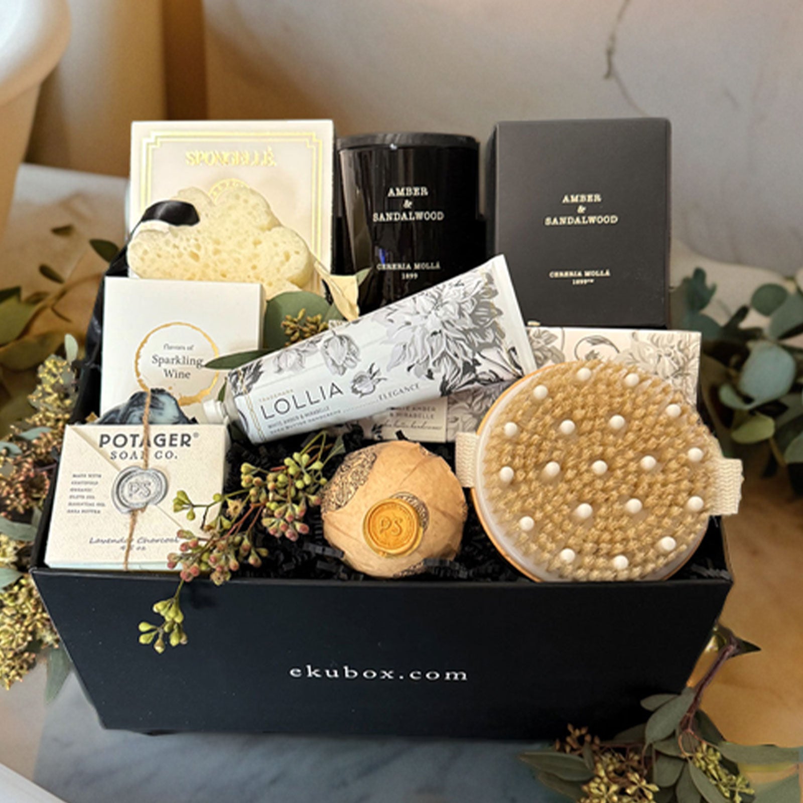 Black pampered spa gift box featuring self-care items including a body wash infused spongelle, cerera molla candle, lollia hand cream, handmade soap, bath bomb, dry body brush, and gourmet gummy treats.
