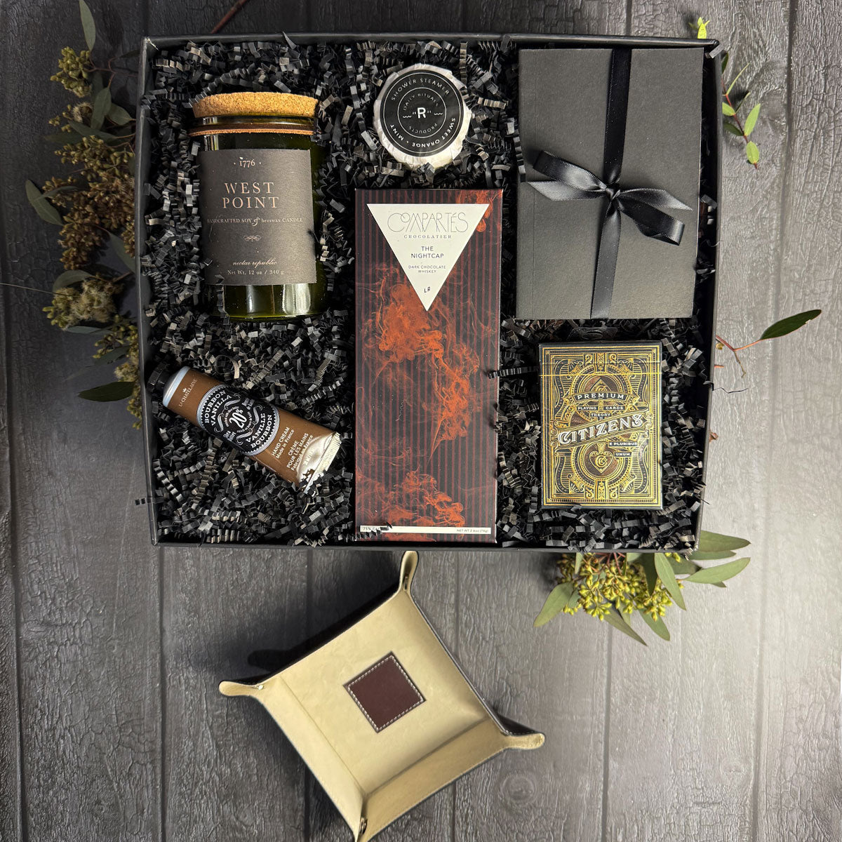luxury gift box featuring a brown leather snap valet, Gunpowder or West Point candle, Theory11 playing cards, chocolate bar, notebook, hand cream, and shower steamer in black box on black background