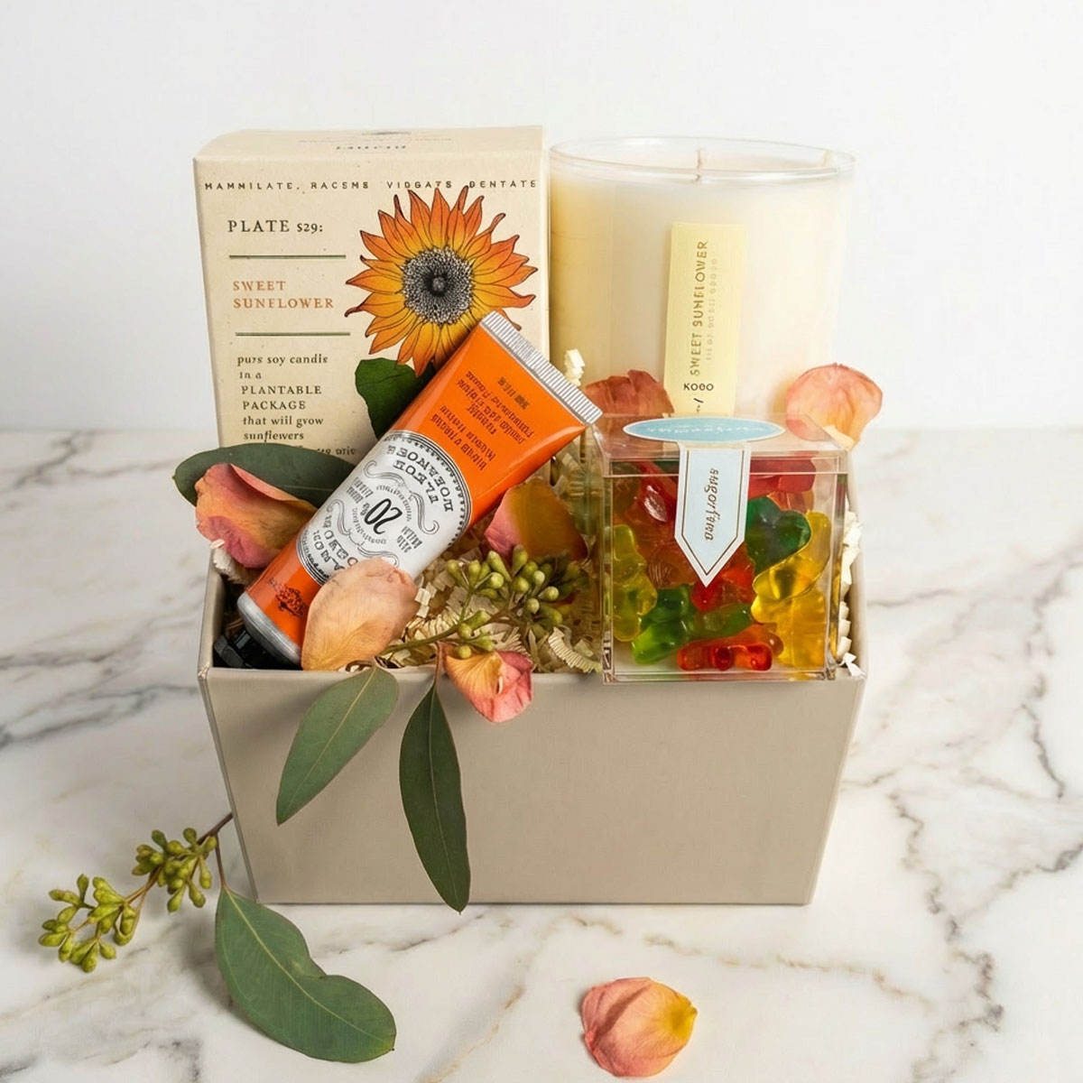 sunflower candle with handcream, sugarfina candy in gift box with rose petals on marble surface.
