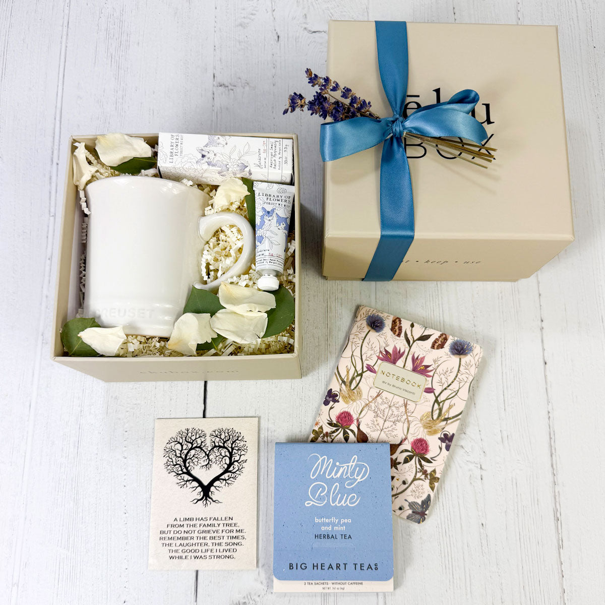 Sympathy gift box with mug, journal, and tea in gift box with ribbon and lavender. Forget me not seed packet on white surface.