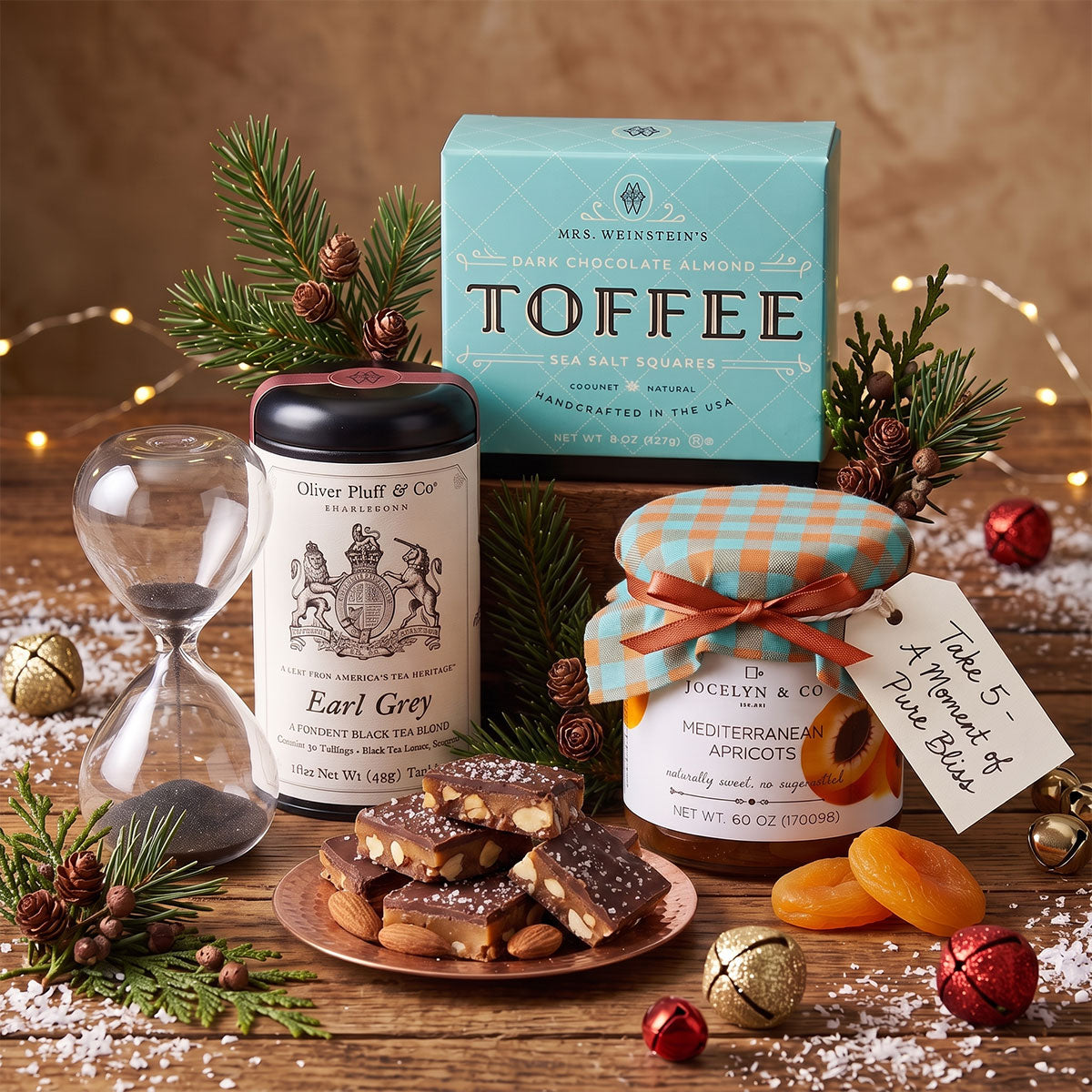 A styled holiday gift set on a wooden surface featuring Mrs. Weinstein's Toffee box, an Oliver Platt & Co Earl Grey tea tin, a jar of apricots with a tag reading 'Table 5- A Moment of Pure Bliss', almonds and dried apricots, pine branches, fairy lights, and an hourglass.