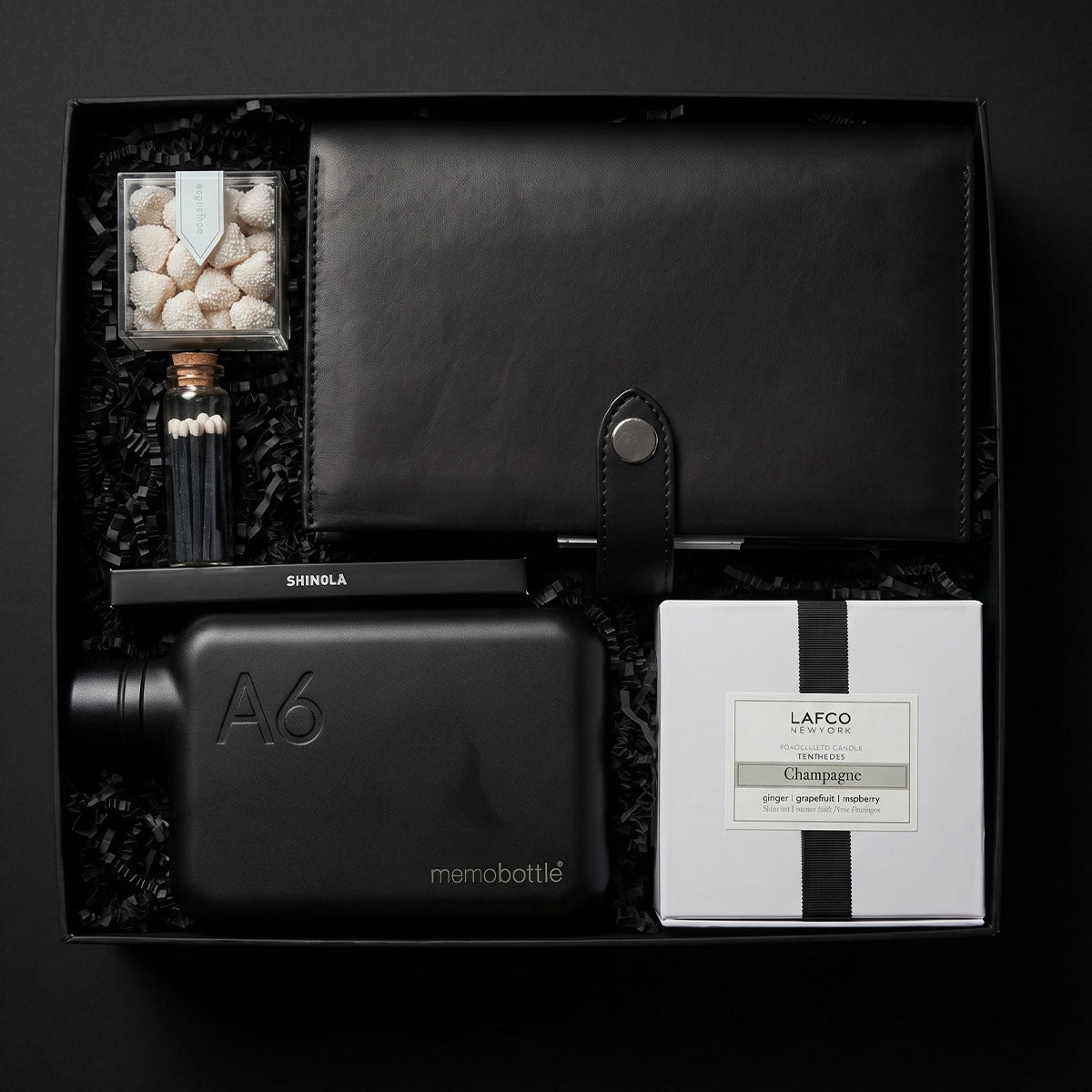 Gift box featuring Shinola notebook cover, shinola notebook, shinola stainless pen, a White candle with black rim and box, a memobottle flask, candies and matches on a dark background
