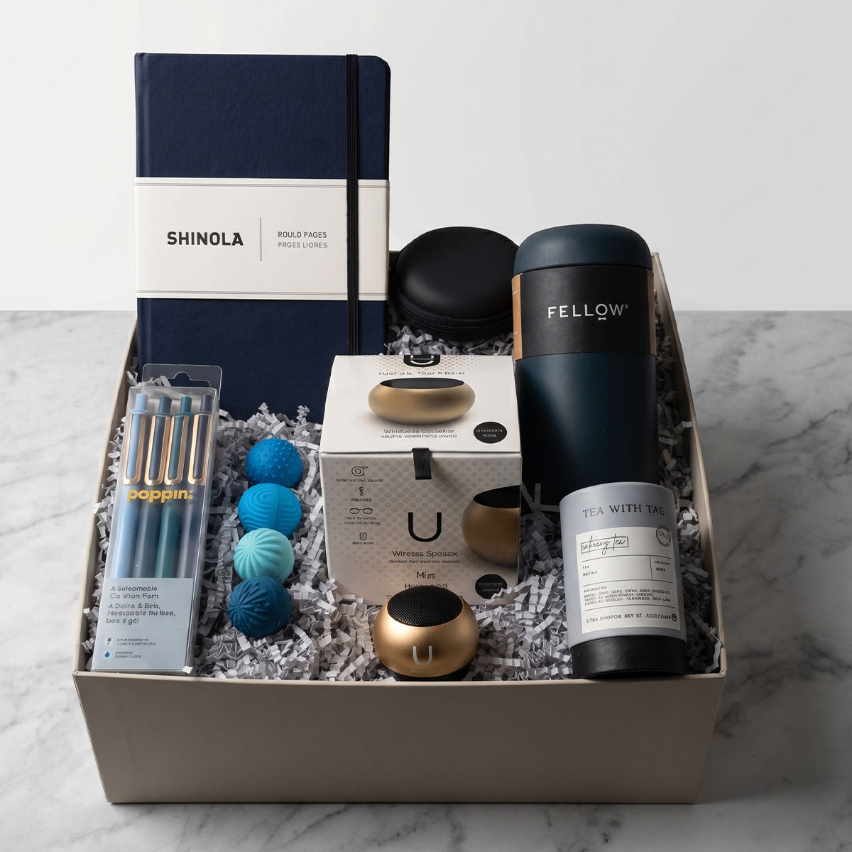 Gift box with Shinola notebook, Fellow tumbler, U Mini Speaker, Poppin Pens, Tea Tube, and 4 stress balls, on a marble surface.