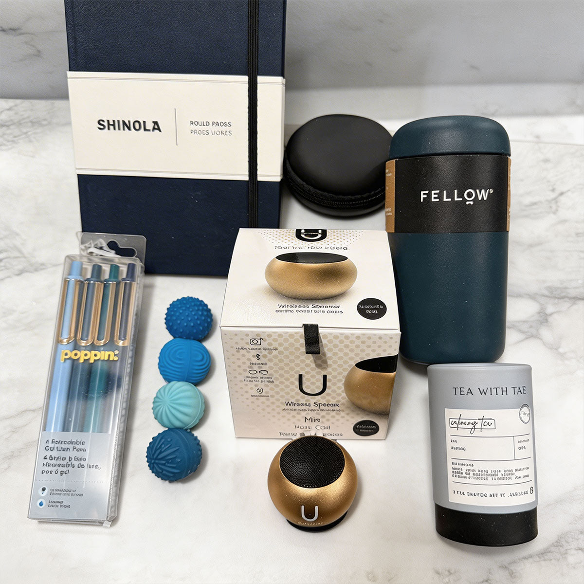 Shinola notebook, Fellow tumbler, U Mini Speaker, Poppin Pens, Tea Tube, and 4 stress balls, on a marble surface.