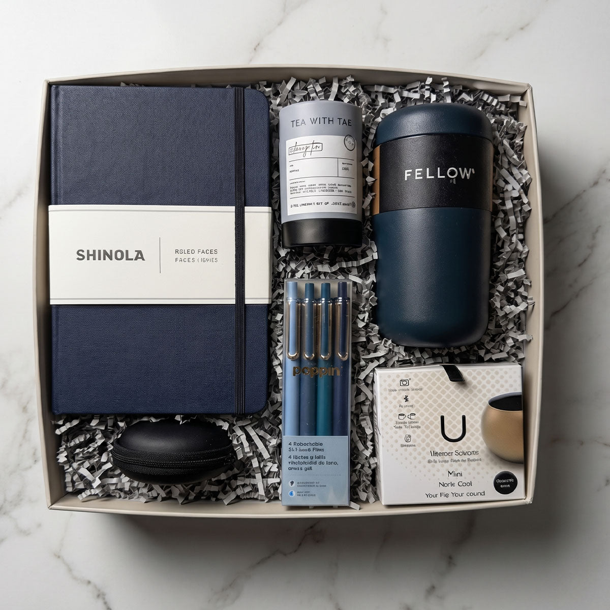 Gift box with Shinola notebook, Fellow tumbler, U Mini Speaker, Poppin Pens, Tea Tube, and 4 stress balls, on a marble surface.