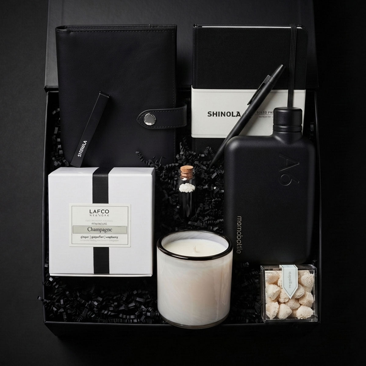 Gift box featuring Shinola notebook cover, shinola notebook, shinola stainless pen, a White candle with black rim and box, a memobottle flask, candies and matches on a dark background
