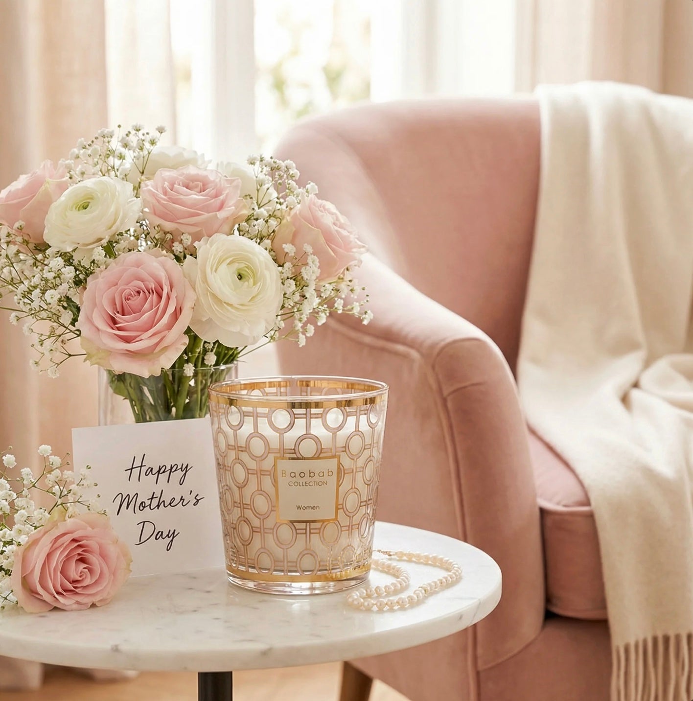 A blush chair with a white throw and flowers on a table, a notecard saying Happy Mother's Day, and a Baobab Women's candle in pink and gold on a small marble tabletop.