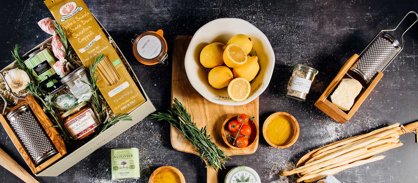 Gourmet food gift box with grissini breadsticks, fresh tomatoes, lemons, a wooden cutting board, cheese grater with Parmesan cheese, and a tin of olive oil.