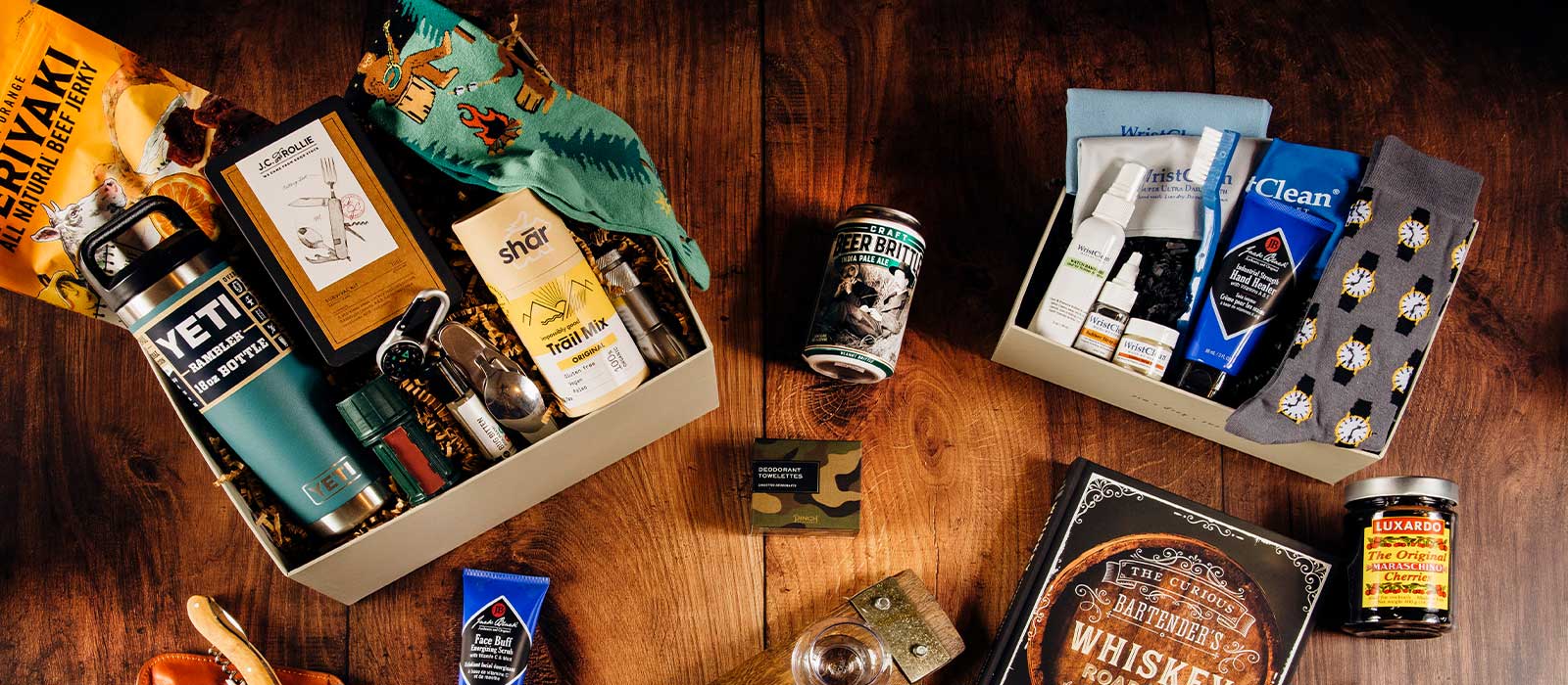 Men’s gift boxes displayed on a wooden surface, featuring a whisky book, socks, and curated gifts for him.