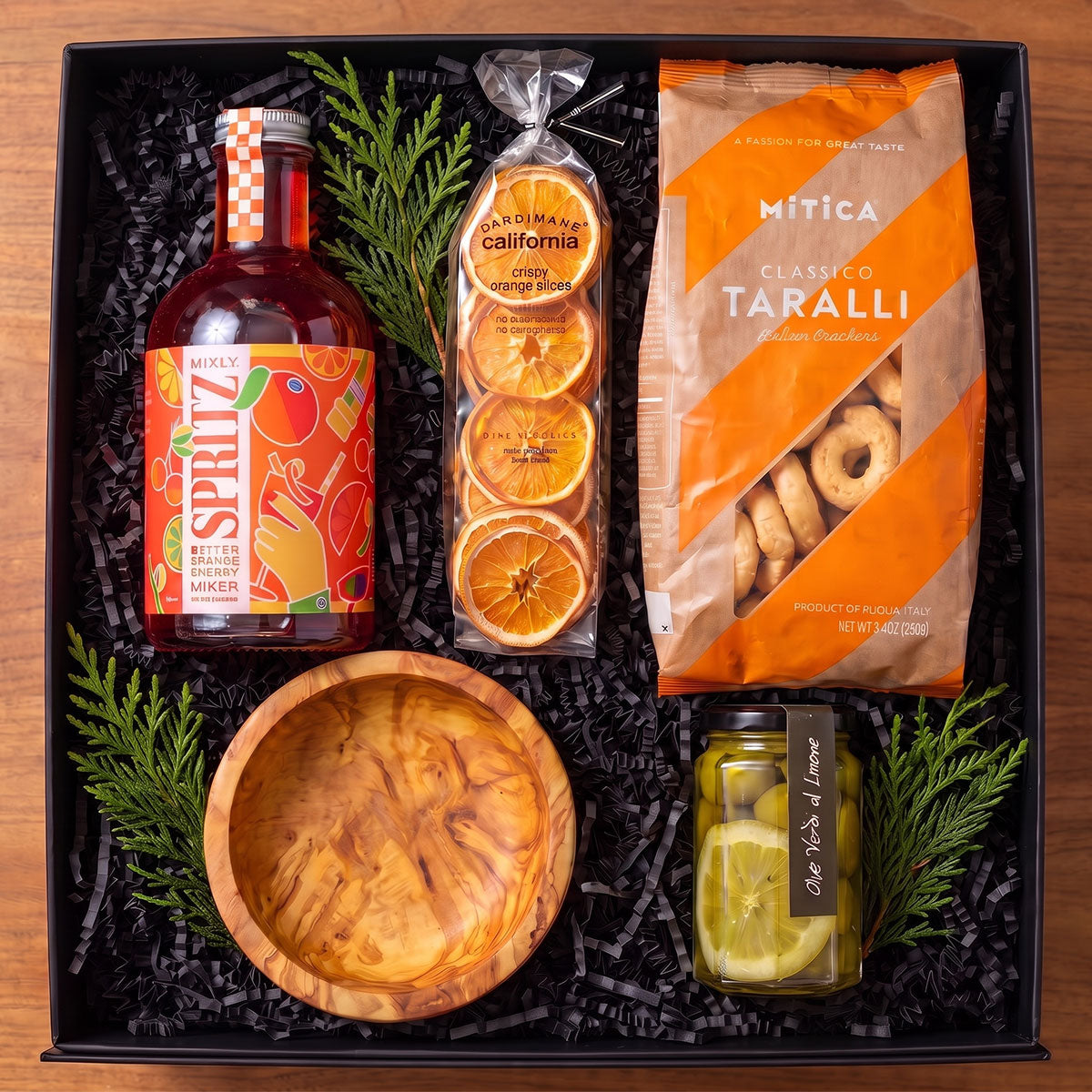 gift box with non-alcoholic Italian Spritz mix, olive wood dip bowl, dried oranges, jar of olives and lemon and taralli bag with holiday greens in the box