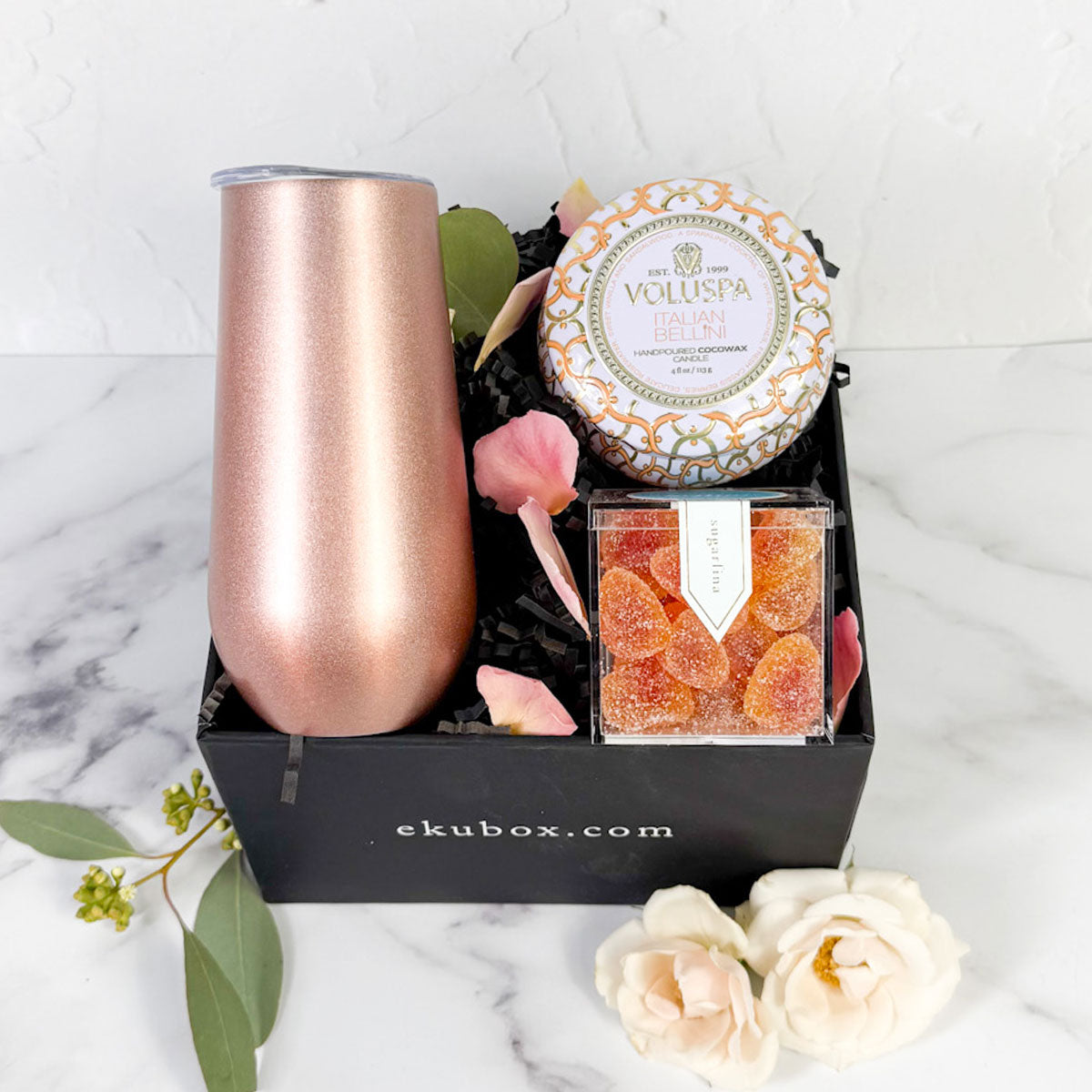 A gift set containing a metallic stemless flute, a Prosecco bellini Voluspa candle, and Peach Bellini gummy hearts in a black ekuBOX with decorative flowers.