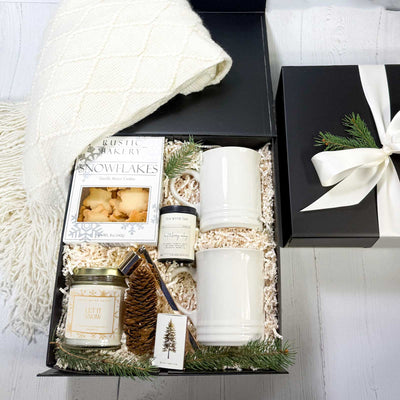 Gift set with a candle, mugs, and a throw blanket, cookies, tea canister,a  candle snuffer, and matches in a black box on a white surface.