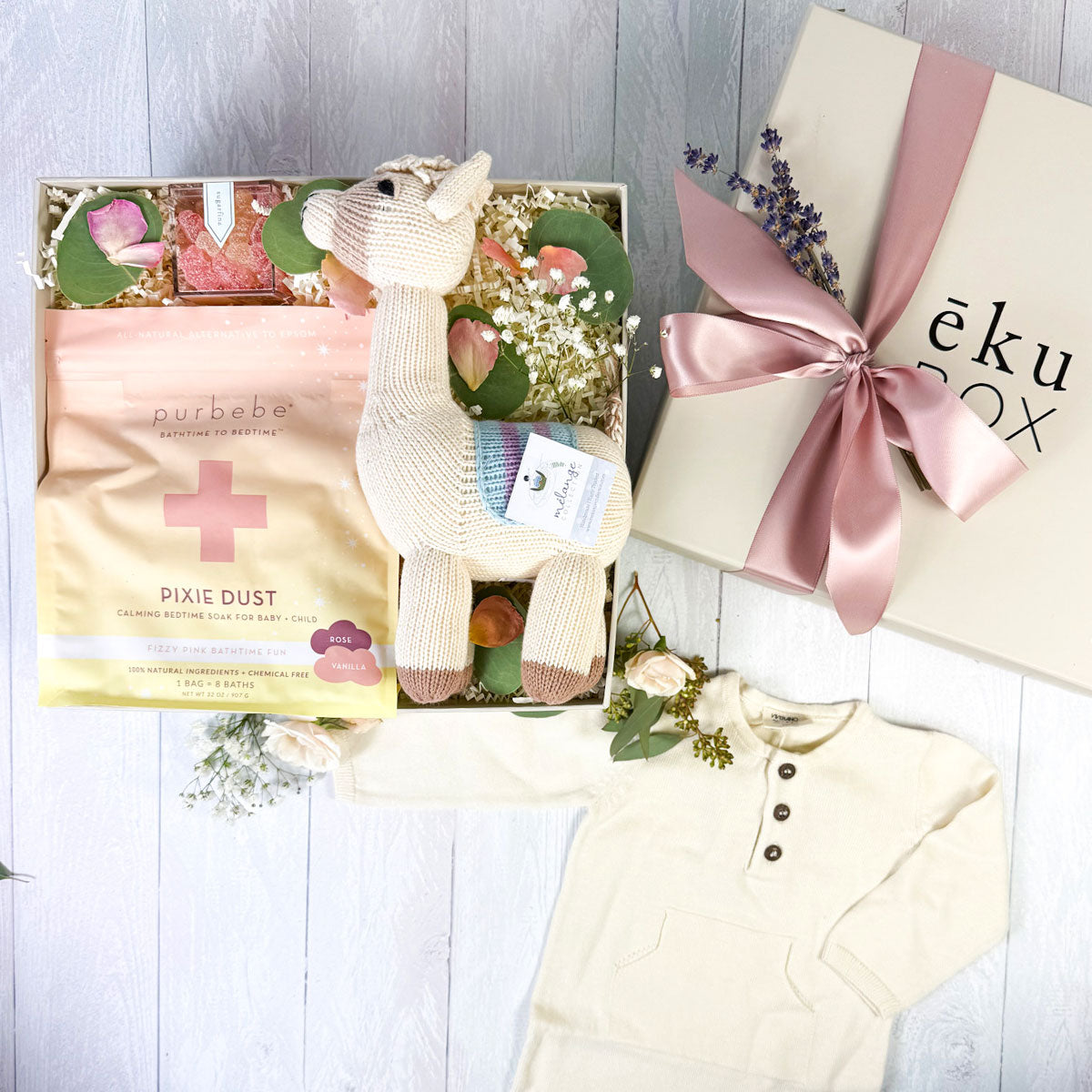 top view of A baby gift box containing a hand-knit cotton llama, an organic cotton kangaroo pocket onesie, a bag labeled 'Pixie Dust', and Sugarfina gummy bears, all arranged in a presentation with eucalyptus leaves and rose petals.