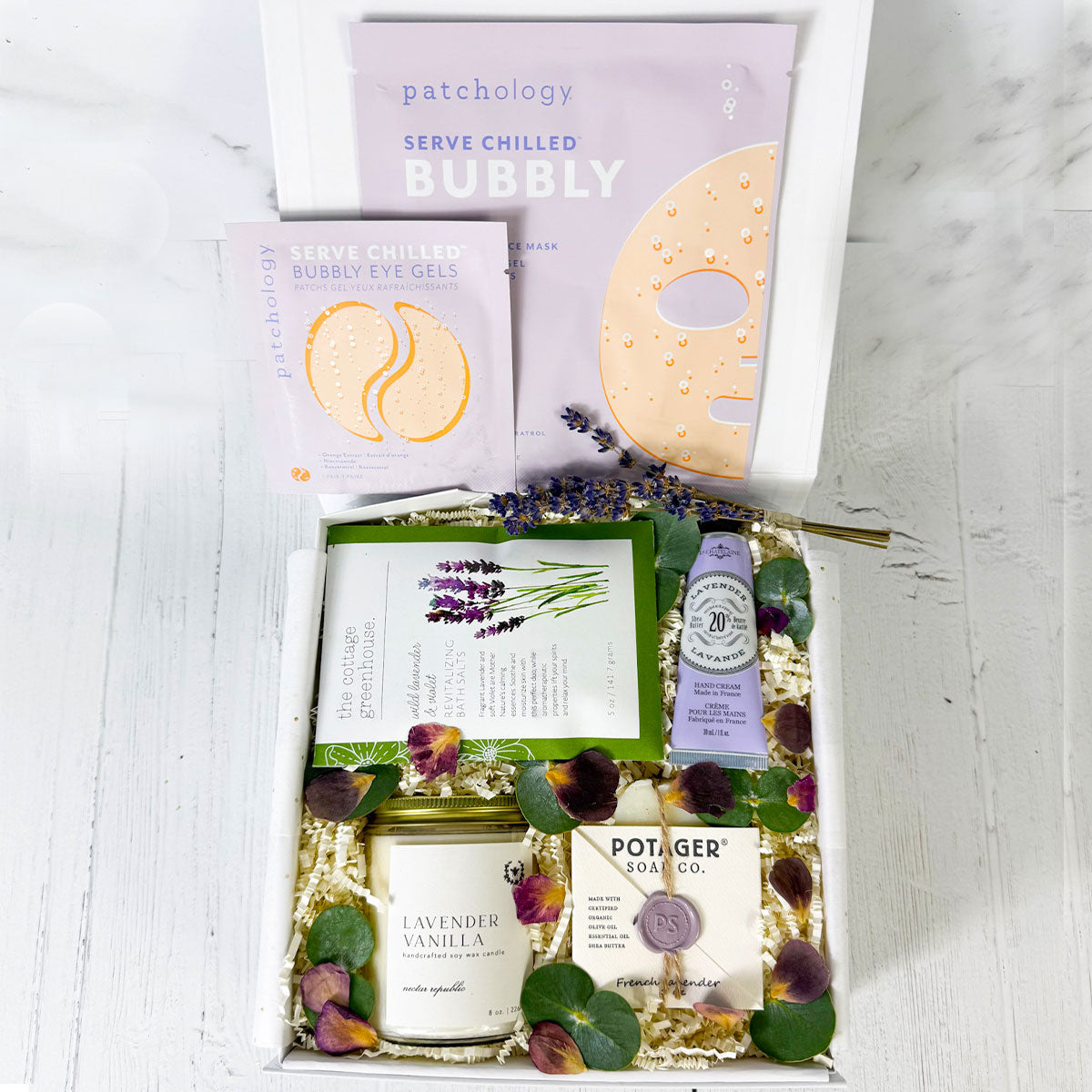 A gift box containing a variety of lavender-themed spa products, including a candle, hand cream, soap, and facial mask, garnished with dried lavender and rose petals.