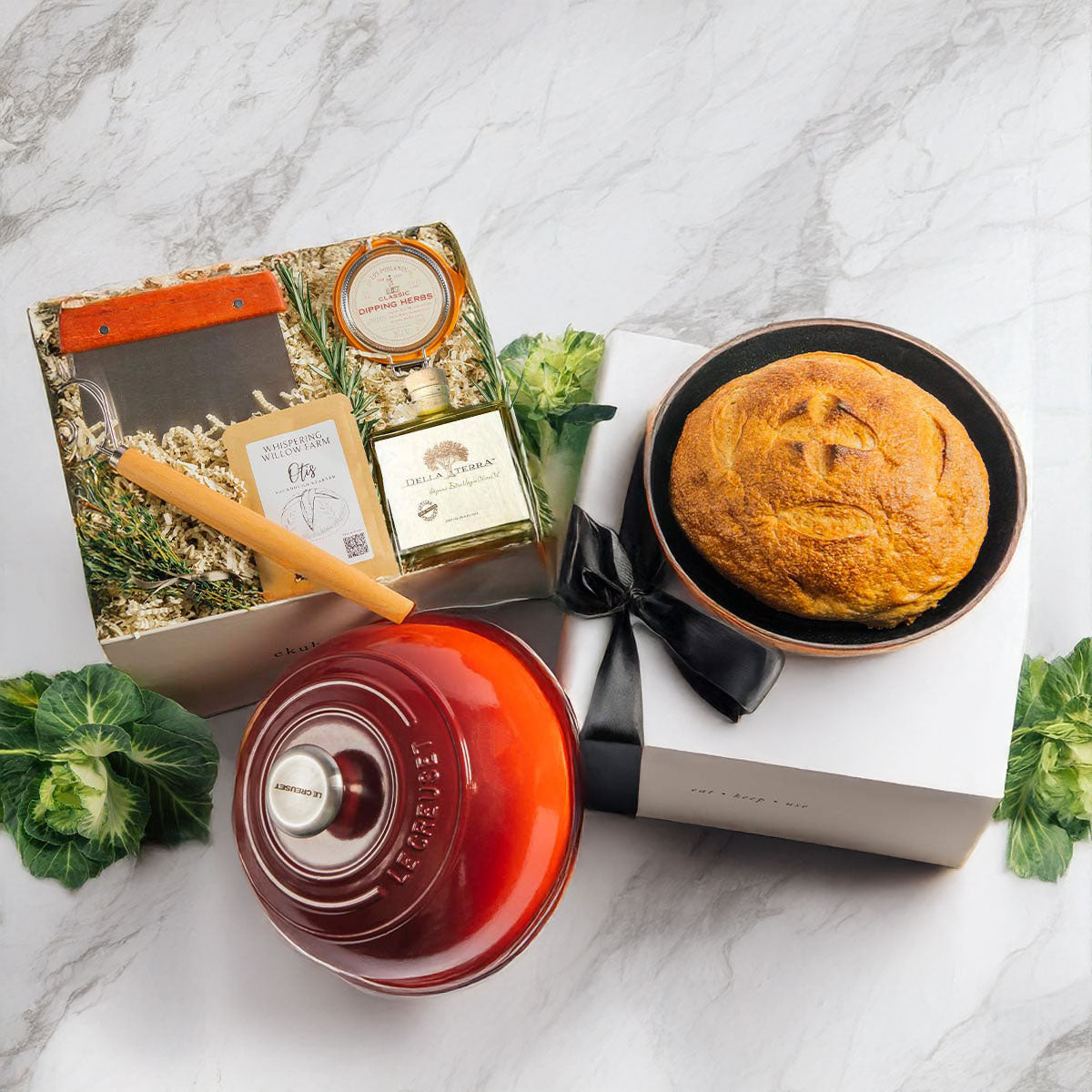 A Le Creuset sourdough bread baking kit with a red bread oven, dutch whisk, sourdough starter, bench scraper, and olive oil with herbs, on a white marble surface.