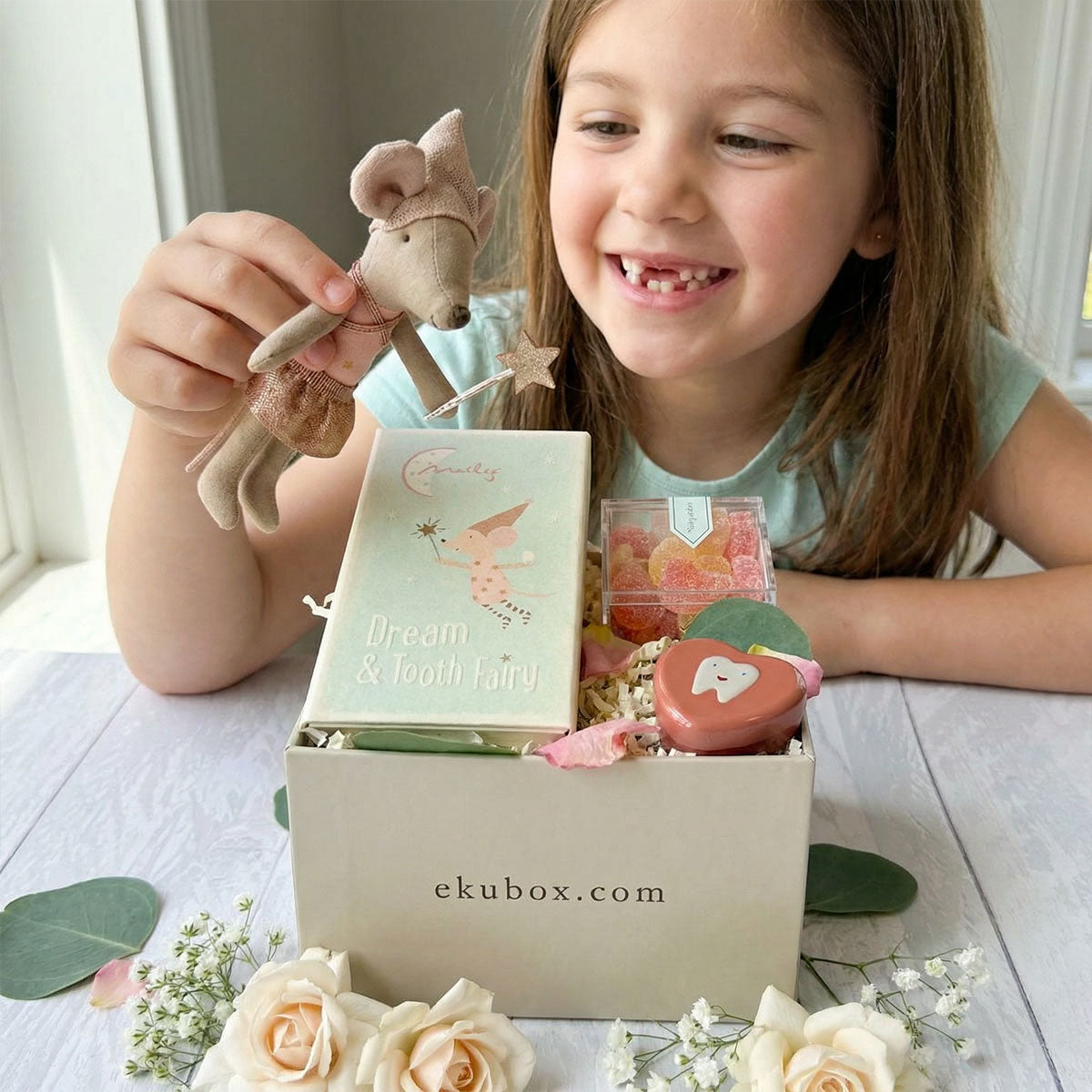 Child holding a Maileg Tooth Fairy Big Sister Mouse with a box of gummies on a table with flowers.