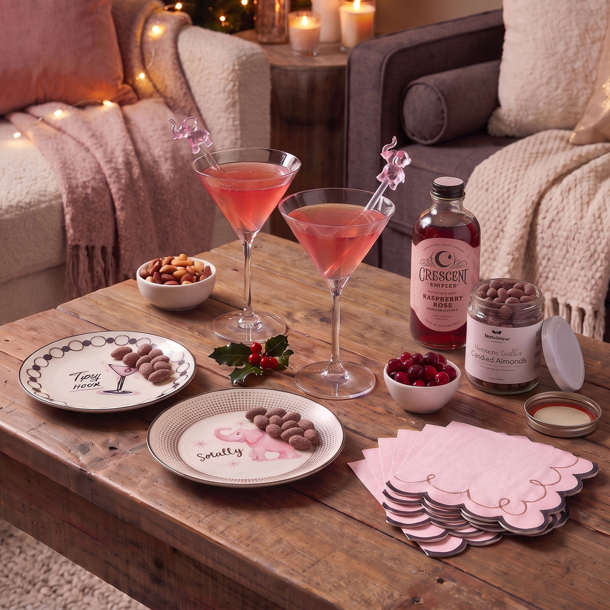 A styled holiday table setting featuring pink and white ceramic plates with scalloped edges, filled with chocolate-covered nuts. Two pink cocktails in highball glasses, a bottle of mixer, a jar of candied nuts and pink napkins are arranged on a rustic wooden table.