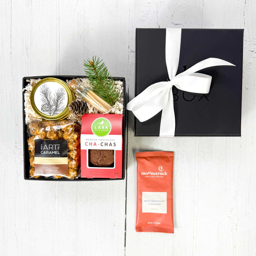 Gift box with popcorn, cha cha cookies, a candle, glass jar of matches, a pine cone a sprig of evergreen, and a black box labeled &