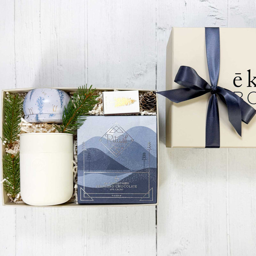 Gift box with candle, box of drinking chocolate, a cream colored ceramic mug, a box of matches and decorative elements on a  white wood surface