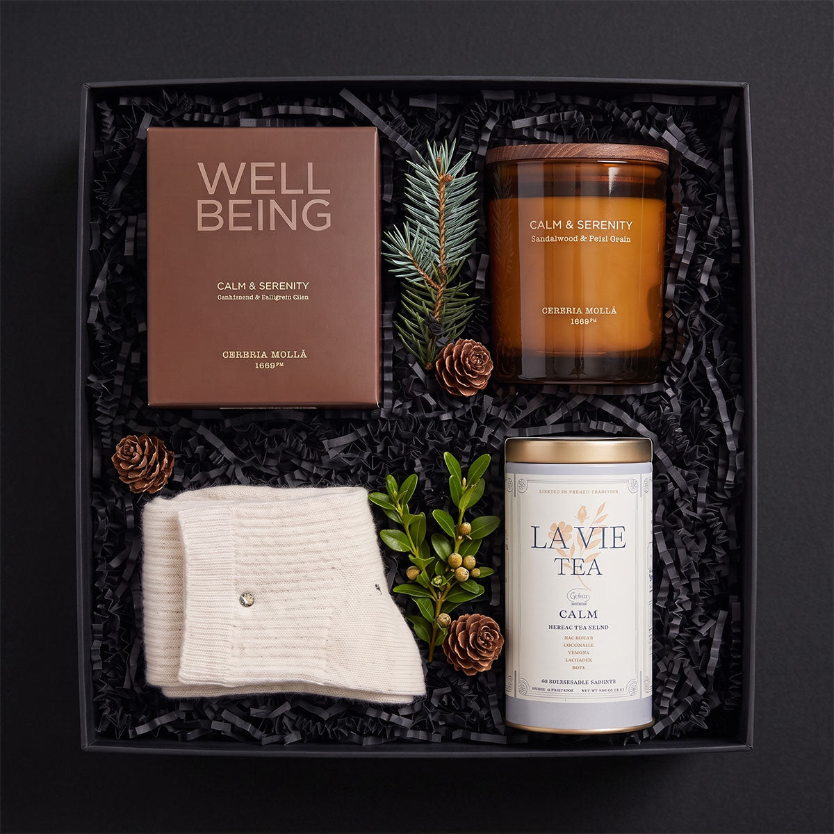 A curated gift set arranged in a black box with shredded black paper filler, featuring a brown Calm & Serenity candle , a pair of chashmere socks and la vie tea clam tea. 