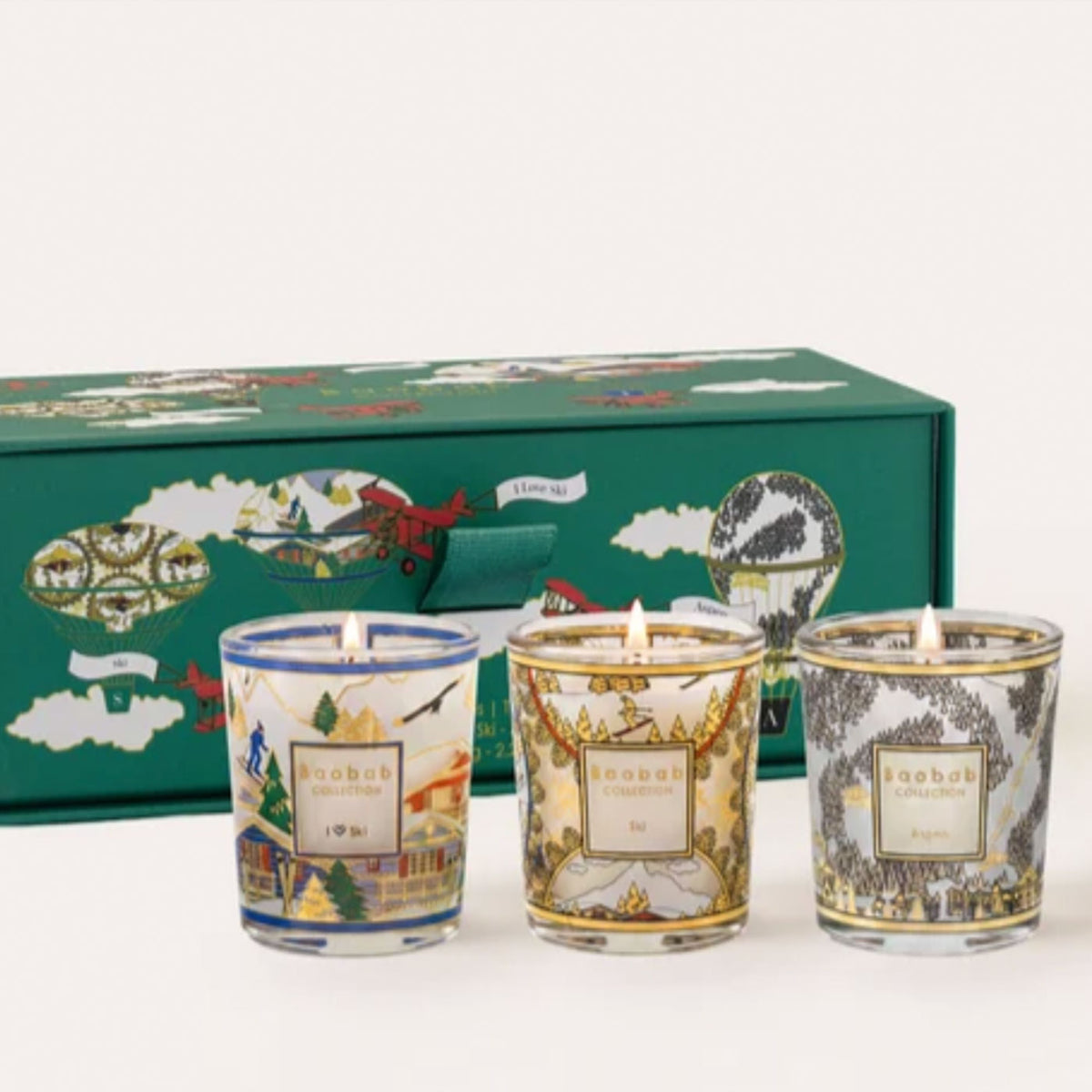 Three small glass scented candles by Baobab with illustrated labels are displayed in front of a green decorative gift box on a white background.