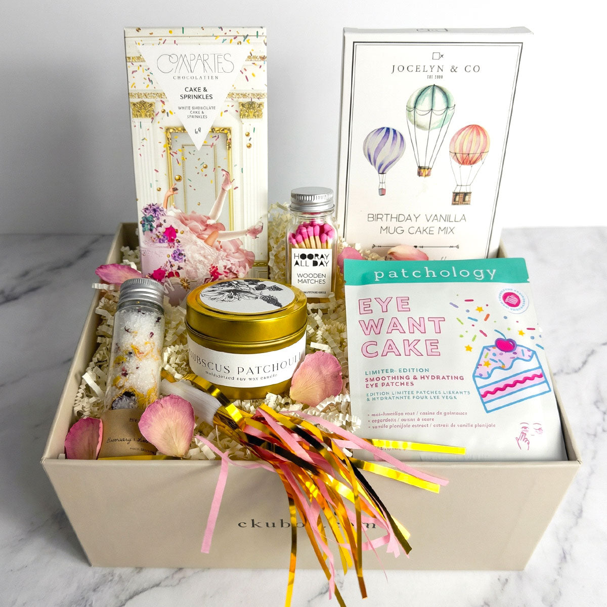 Birthday gift box featuring cake-themed treats and self-care items, including a vanilla mug cake mix, sprinkle chocolate bar, candle, colorful matches, eye patches, floral bath salts