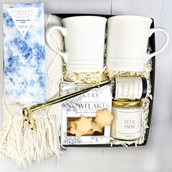 Cozy Moments Gift set with two Le Creuset mugs, a Let it snow candle, a box of snowflake cookies, tea, a throw blanket, a candle snuffer, and a gourmet chocolate bar in a box on a white background